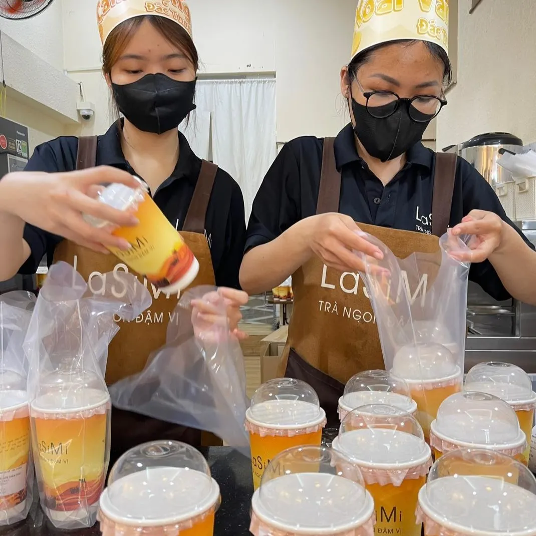 Trà Sữa LASIMI Trà Ngon Đậm Vị Uống Là Nghiền 2 Nhân viên LaSiMi trong tạp dề nâu in logo Trà Ngon Đậm Vị đang đóng gói đồ uống